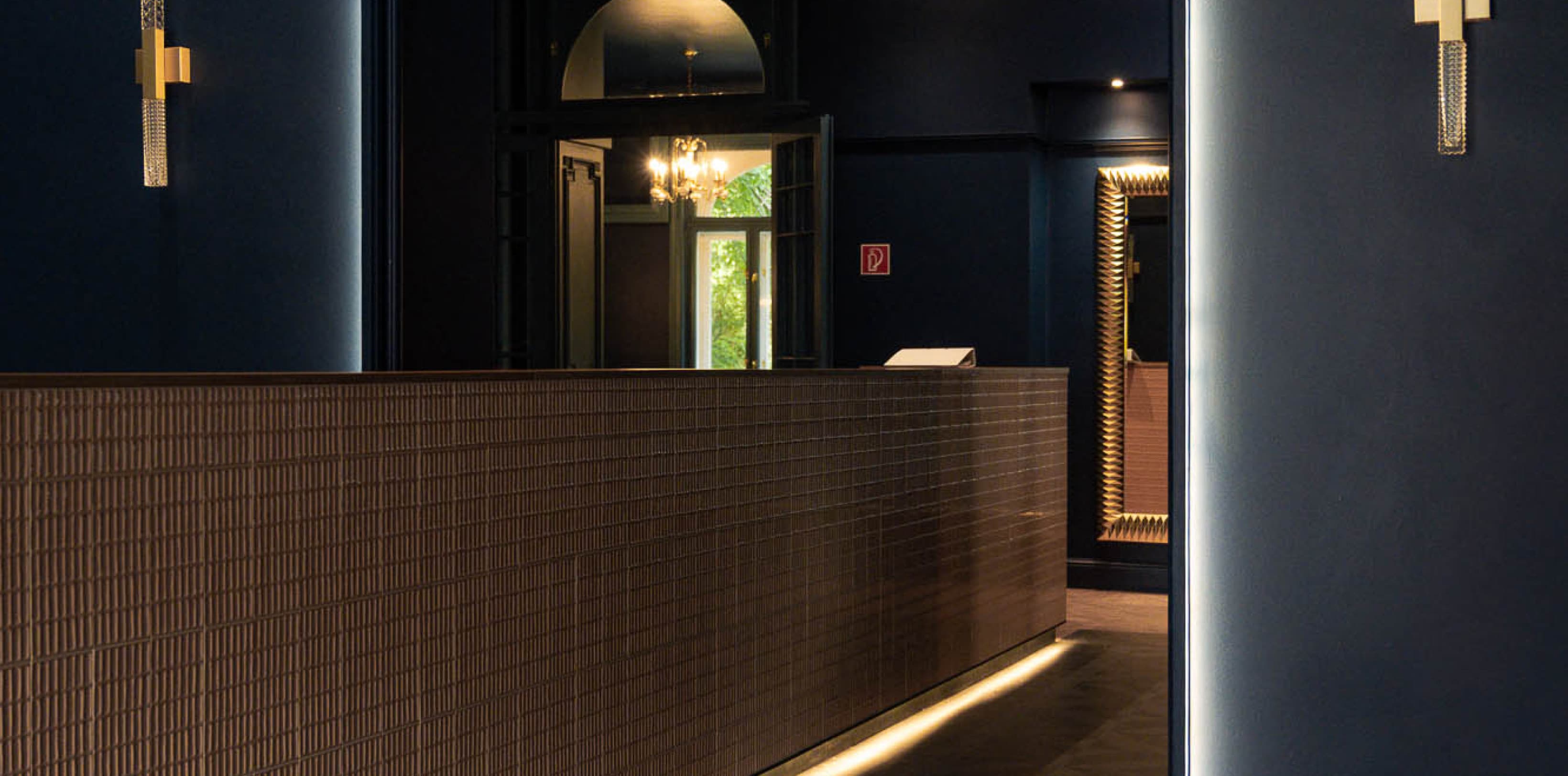 Zu sehen ist ein Torbogen, der sofort ins Auge fällt, wenn man das Hotel betritt. Das Dunkelblau wird durch ein helleres Gelb ergänzt, das an einer barartigen Wand zu sehen ist, die sich bis zum Ende der Lobby erstreckt.